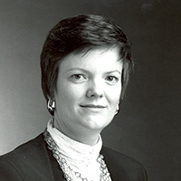 Black and white headshot of Kathleen G. Murray. She is a white woman with short brown hair. She is wearing a black blazer and white turtle neck with a layered chain necklace.