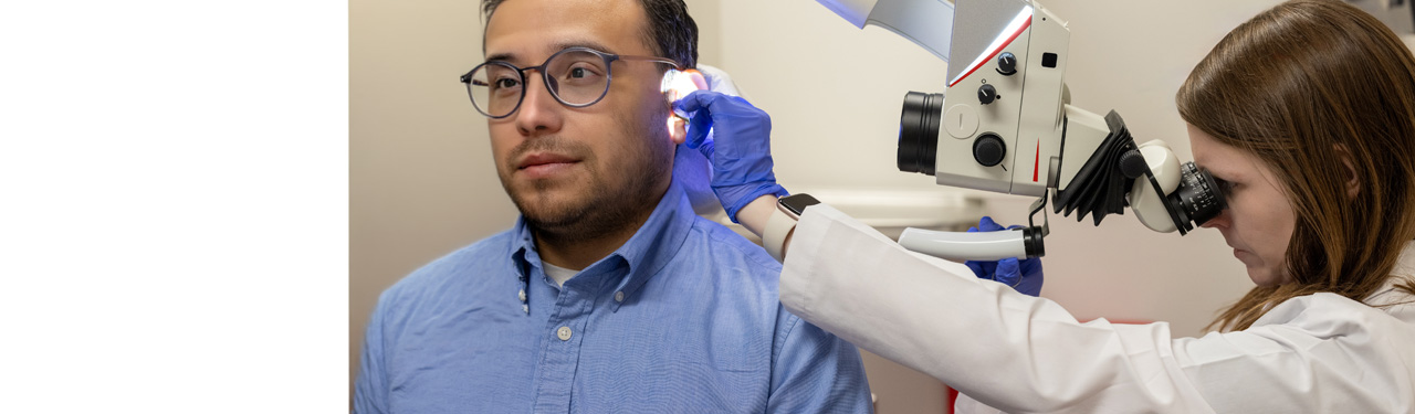 A man wearing a blue shirt and glasses having his inner ear examined through a microscope by a physician.
