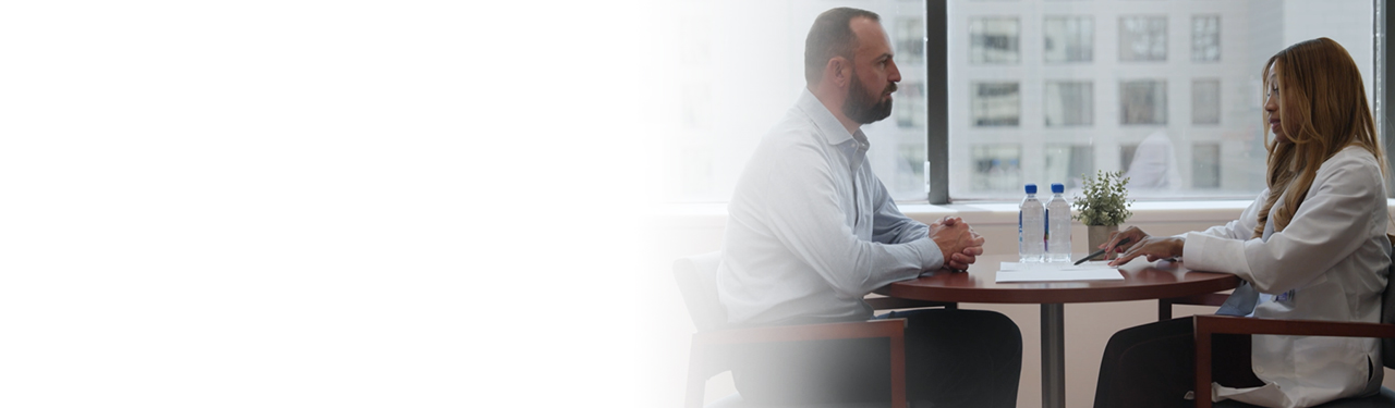A female provider wearing a white lab coat sitting at a table and talking to a middle age man.