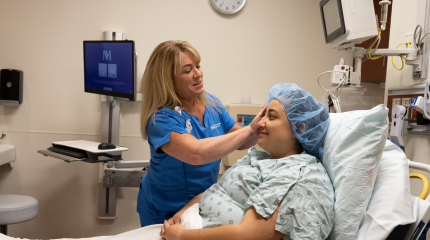 A female nurse wearing blue scrubs helping a female patient on a hospital bed put on a surgical hair net.