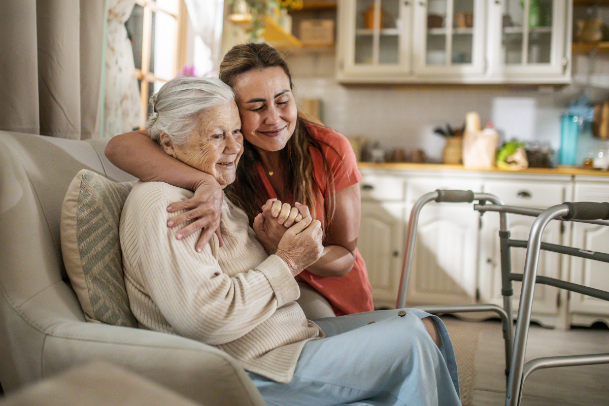 Older adult seated at home holds hands with a caregiver; a walker is nearby.