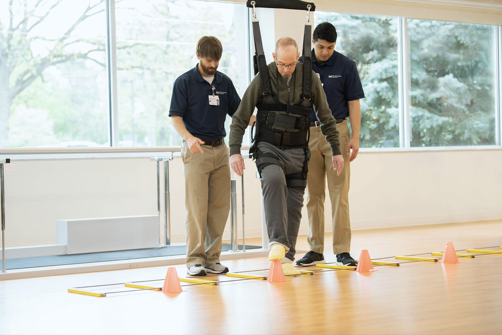 Clinicians helping patient to walk using assistive device suspended from ceiling.
