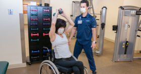 Male clinician standing next to a female patient. She is in a wheelchair and is raising a weight over her head.