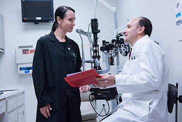 A male Ophthalmologist wearing a white lab coat talking with a female employee wearing an all black outfit.