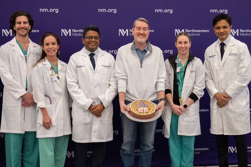 Surgeons stand in front of a purple wall. Their patient is holding a plate with a large cake/cookie on it.