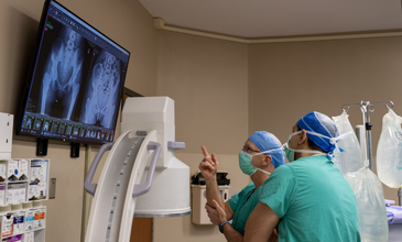 Two male physicians wearing green scrubs and face masks pointing to an X-ray of a patient's hips on a TV screen.