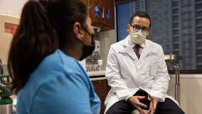A physician in a white lab coat and yellow surgical mask sitting and talking with a hispanic woman wearing a blue shirt and black mask.