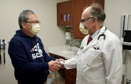 A male Northwestern Medicine physician wearing a yellow surgical mask and white lab coat shaking the hand of a male Asian patient wearing a blue sweatshirt and yellow surgical mask.
