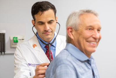 Doctor Seides with the Northwestern Medicine Pulmonology team listening to the heartbeat of an elderly male patient who is smiling.