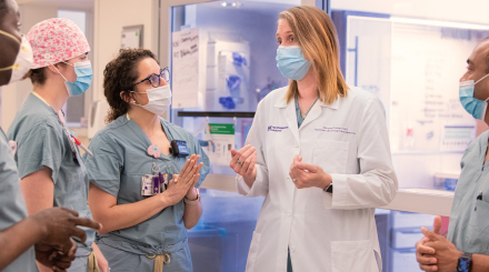 A tall blonde hair female physician wearing a white lab jacket talks with a team of nurses who are all wearing blue scrubs and surgical masks.