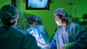 Three physicians in blue scrubs standing in a green lit operating room under operating lights.