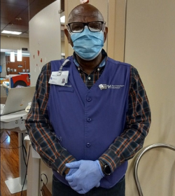 An African American elderly man wearing a purple vest and mask looking at the camera.