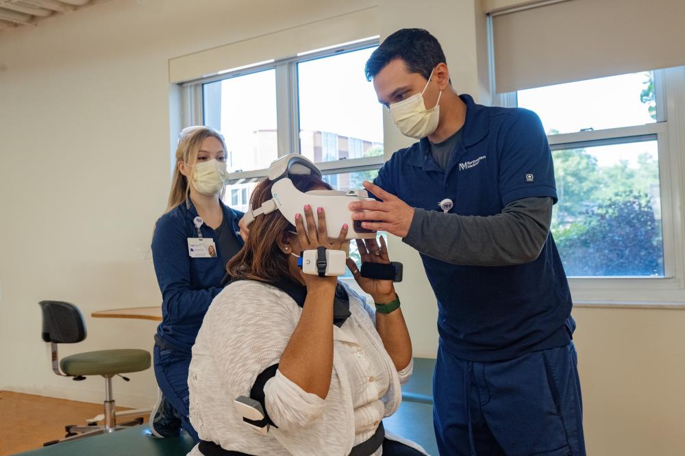 Male physical therapist in a mask helping a woman use a virtual reality headset.