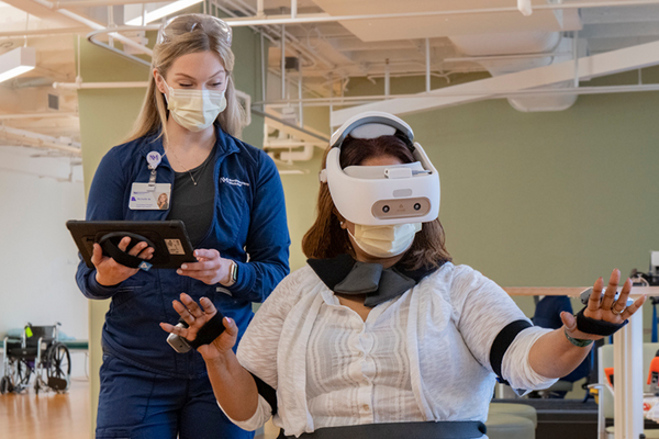 Female patient wearing a VR headset. Female physical therapist in an NM jacket is holding a tablet and assisting her.