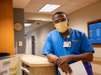 A male nurse wearing blue scrubs, a surgical face mask and medical glasses leaning against a counter.