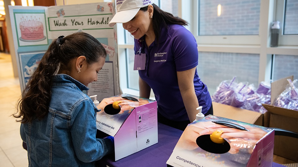 A Northwestern Medicine employee wearing a purple shirt and white hat talking to a young girl at a community event booth.
