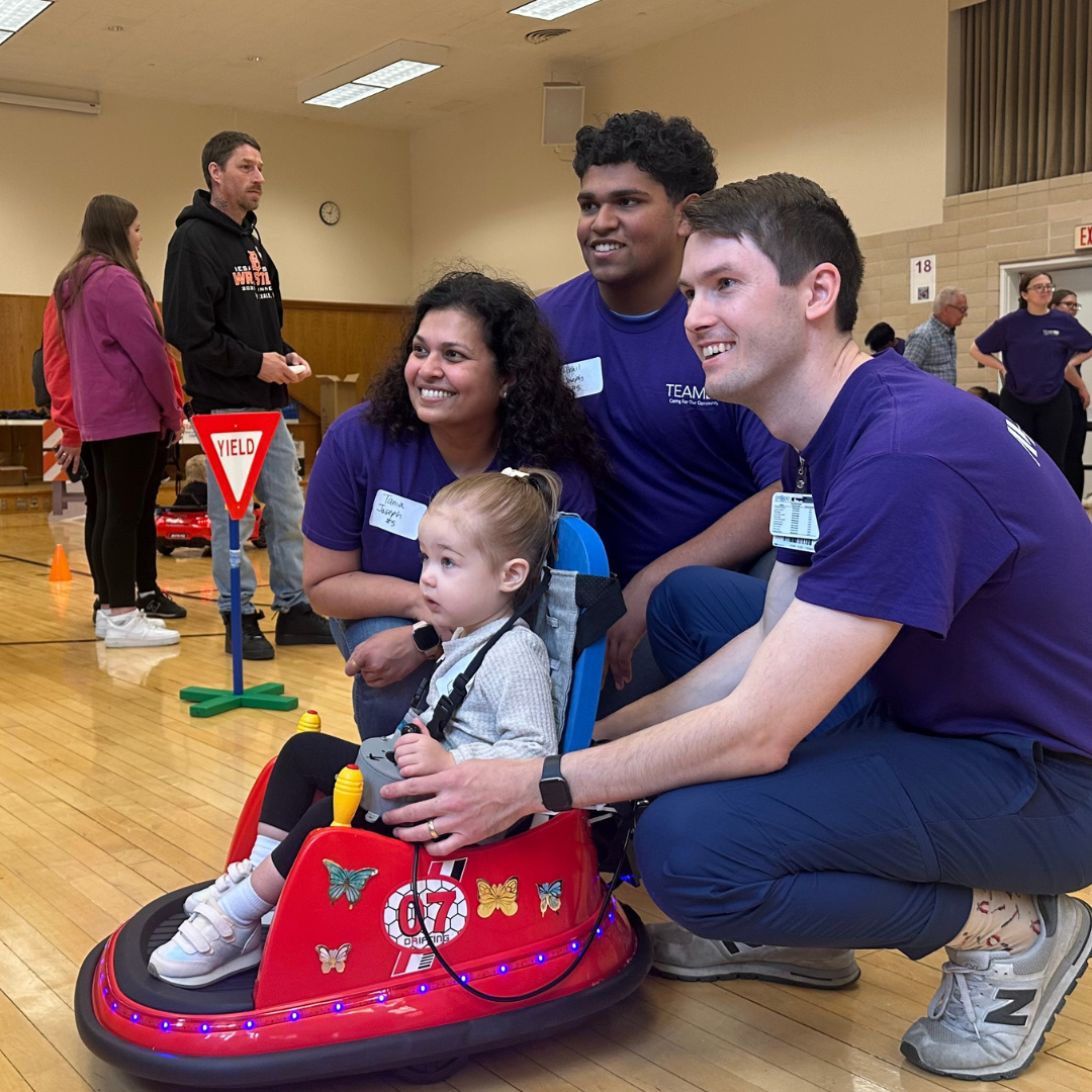 Northwestern Medicine team members interact with a child seated in a modified ride-on toy car at a community event.