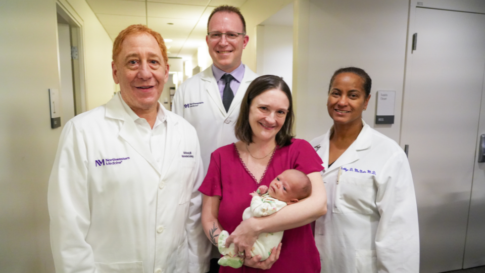 Patient holds a newborn infant while standing with members of the care team in a hospital hallway.
