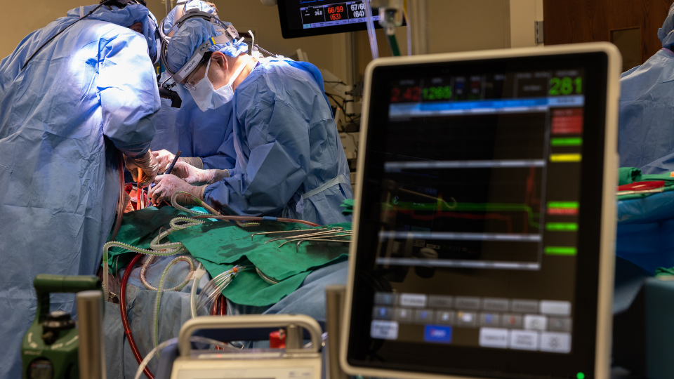 Surgical team performs a cardiac procedure in an operating room with patient monitoring equipment visible.