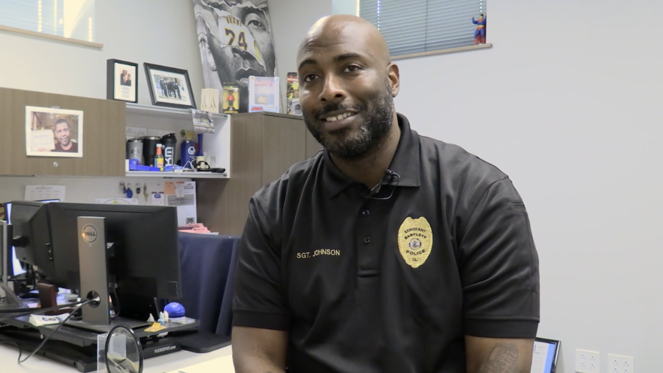 Police sergeant sitting behind a desk, wearing a shirt with his name and badge.