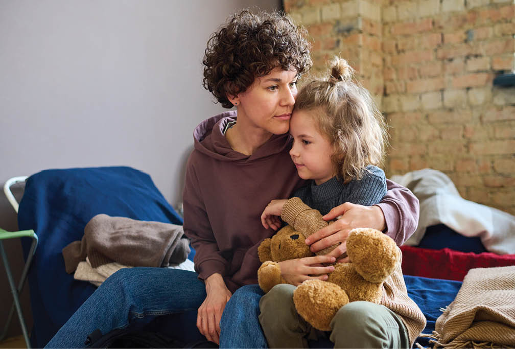 Young woman giving hug to her cute little son with brown soft teddybear while both sitting on sleeping place prepared for refugees