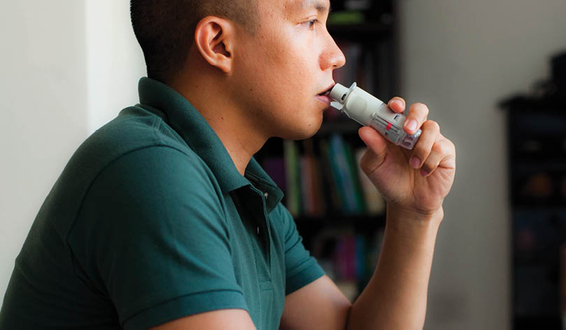 Side view portrait of a young Southeast Asian man using an asthma inhaler device indoor