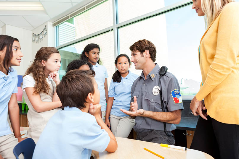 A diverse group of school children are interested in what a police officer has to say about safety in their school. They are wearing school uniforms. Their teacher is standing next to the officer.