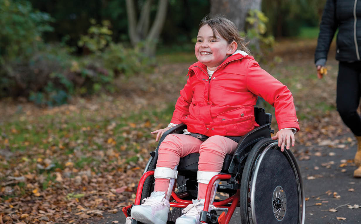 A low-angle view of a young girl out with her family for a day out in a public park in Newcastle upon Tyne. She is a wheelchair user and is pushing herself ahead of her family in the park.