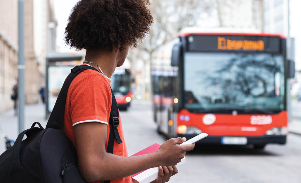 Side view of unrecognizable young African American man in casual clothes and backpack using smartphone while standing on city street and waiting for bus