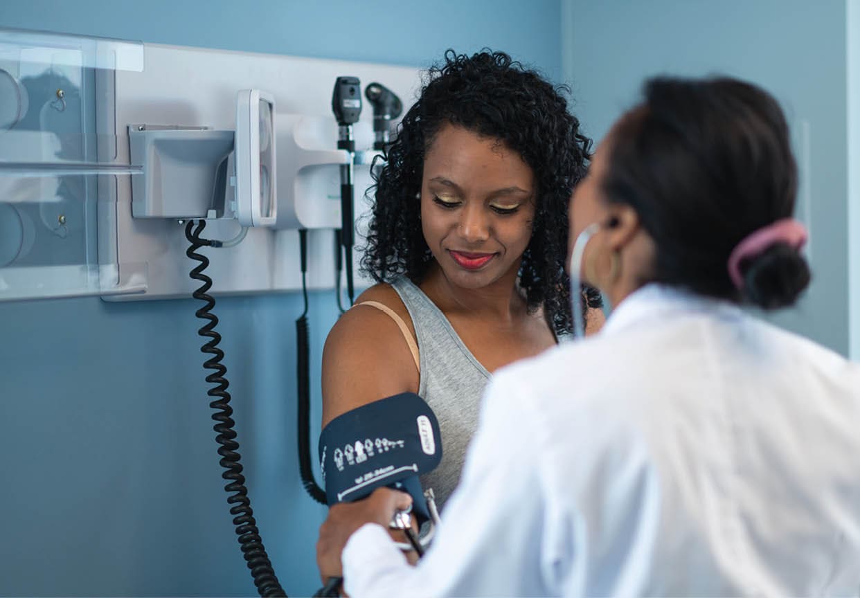 A young black woman is at a routine medical appointment. Her healthcare provider is an ethnic woman. The patient is sitting on an examination table in a clinic. The doctor is checking the patient's blood pressure. The patient is smiling while looking down at her arm.