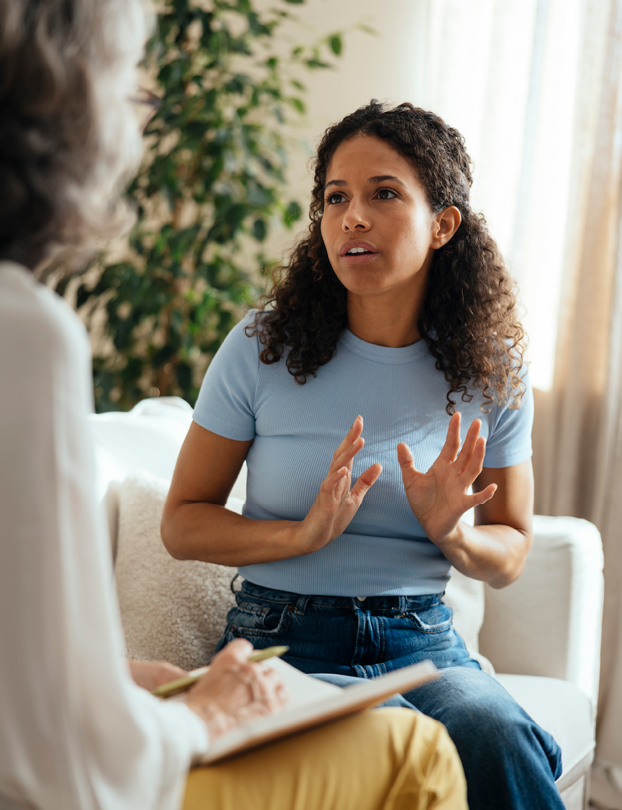 A person with curly hair wearing a light blue shirt and jeans is sitting on a couch and speaking while another person is taking notes on a clipboard. Description generated by AI