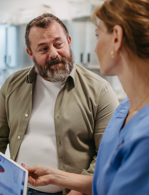 An adult with a beard wearing a green shirt is attentively listening to another adult in blue scrubs who is holding a tablet. Description generated by AI
