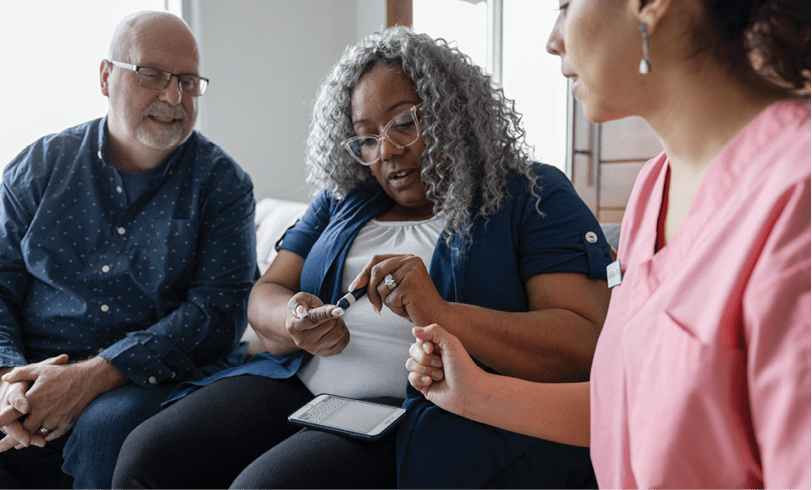 Home caregiver teaching senior woman how to use glaucometer on living room sofa at home