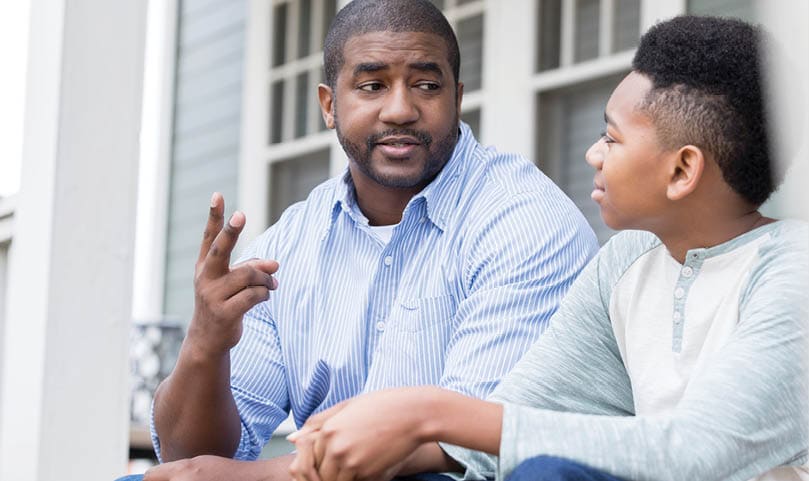 A serious mid adult father sits on his front porch steps with his preteen son. He holds up two fingers for his son as he numbers each consequence for his bad behavior.