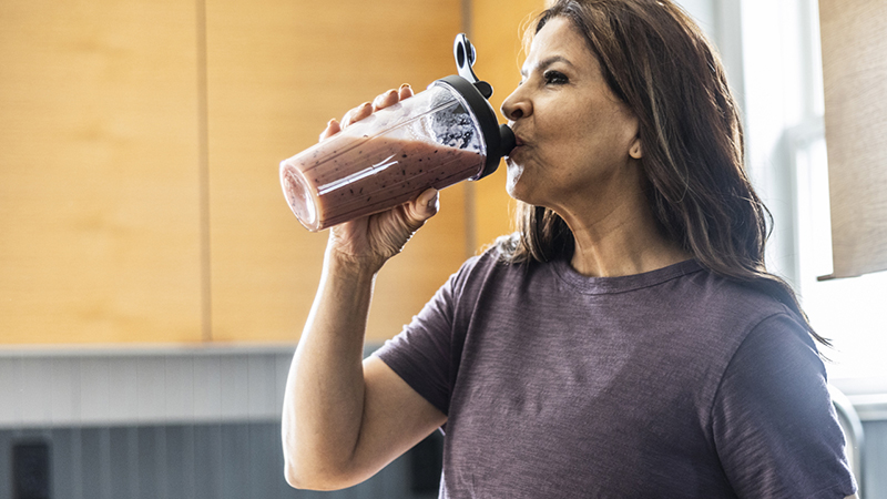 A person with long brown hair wearing a purple short-sleeved shirt drinks a protein shake in the kitchen with supplements and vitamins on the counter. 