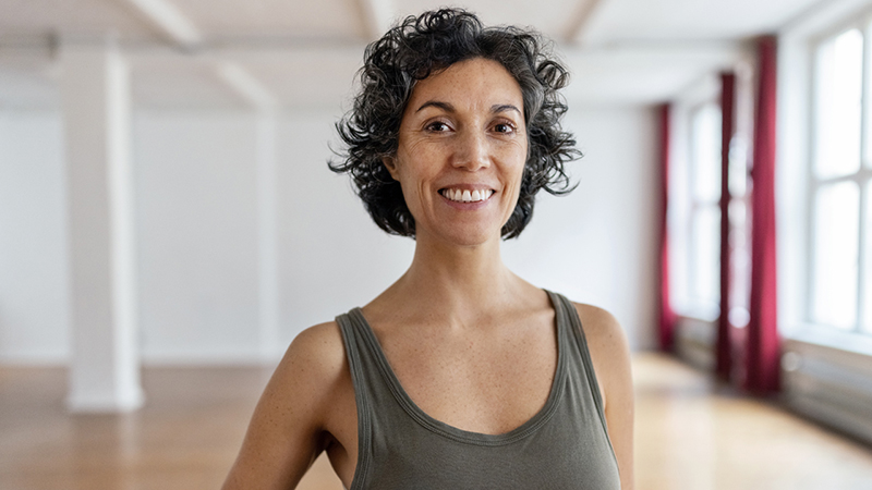 Mature woman with dark, curly hair, brown eyes and freckles wearing a tank top in a yoga studio with wood floors and tall windows in the background.