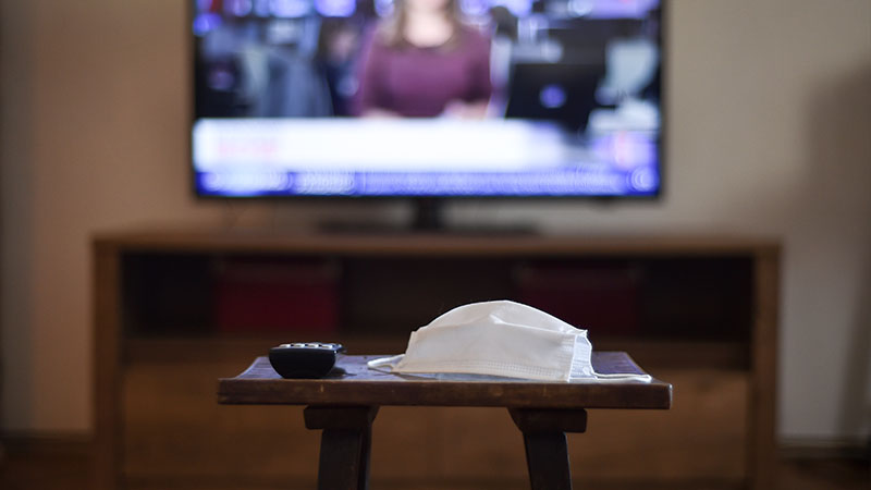 television remote control and a medical mask, with news channel open in background