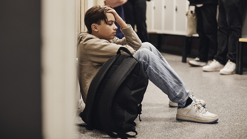 Teen sitting on floor of school hallway with a backpack and head in hand.