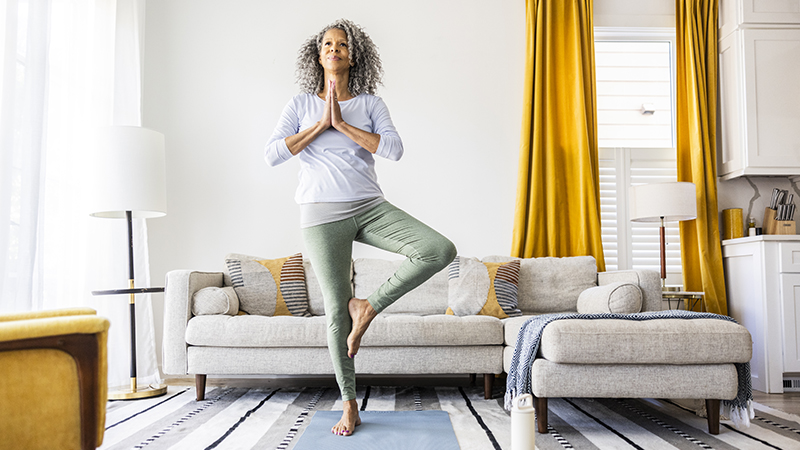 A person with long gray curly hair practices Tree Pose on a yoga mat in a living room. 