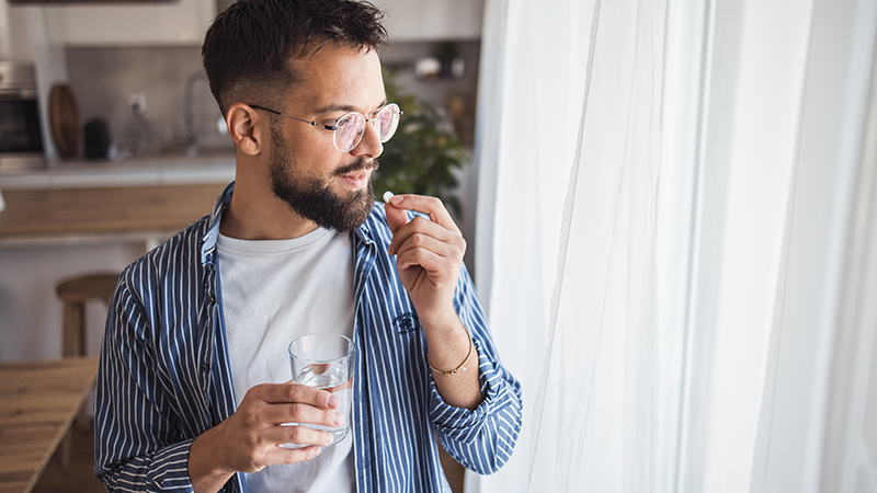 A person stands getting ready to take medication while holding a glass of water and a pill.