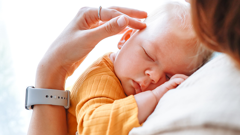 Adult with long brown hair wearing a white sweater and flexing their wrist, illustrating symptoms of mommy’s thumb or carpal tunnel syndrome, while holding a baby wearing a yellow outfit