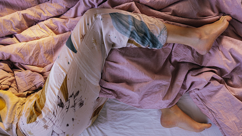 Person laying in bed pictured from the waist down with a lavender colored comforter.