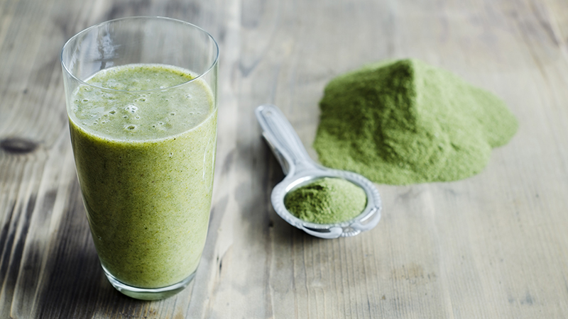 Green smoothie in a glass with green powder and a stainless steel spoon next to it on a wooden table. 