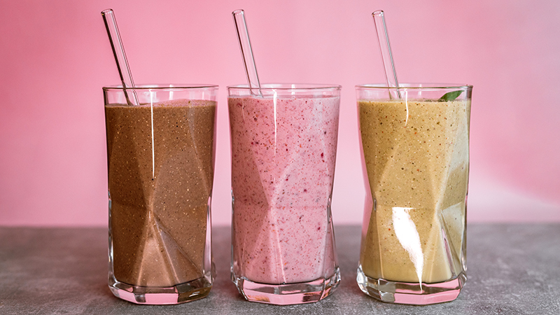 Three different smoothies in glasses with glass straws with a pink background. 