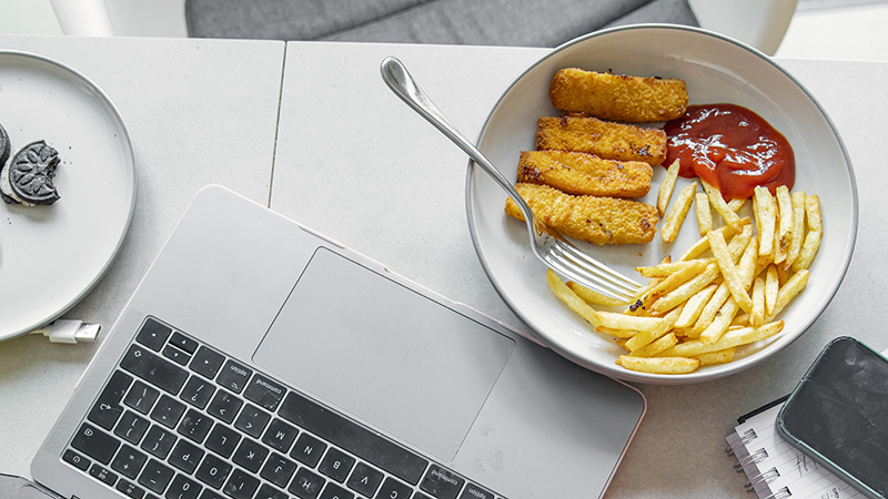 Ultra-processed foods on two plates, including cookies and French fries, fish sticks and ketchup with a glass of orange juice at a desk workstation