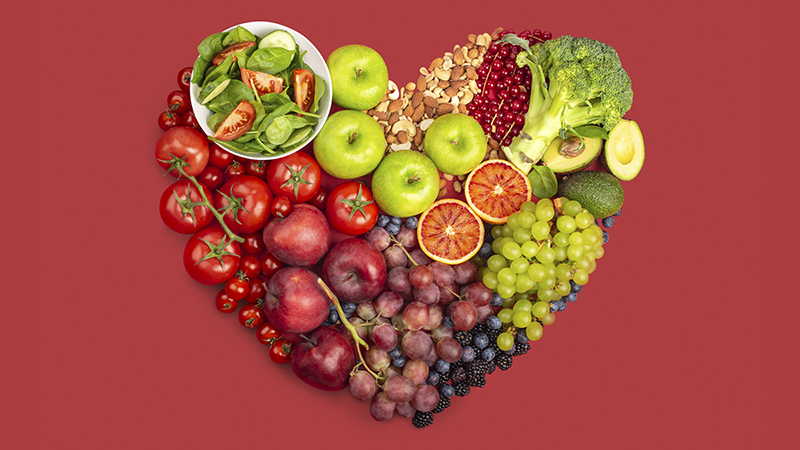 Several fruits, vegetables and nuts in the shape of a heart on a red background, representing healthy eating for heart health.