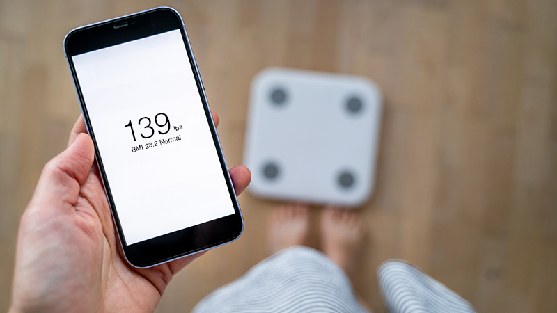 Person standing in front of a scale and holding a smart phone showing their weight and body mass index 