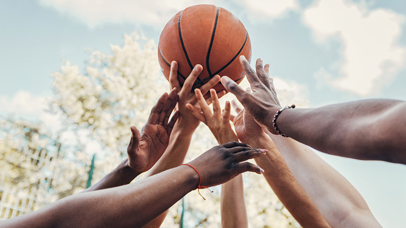 Seven hands of varying skin tones reaching for a basketball at tip off against a washed out blue sky with white clouds. A tree and gate are in the background.
