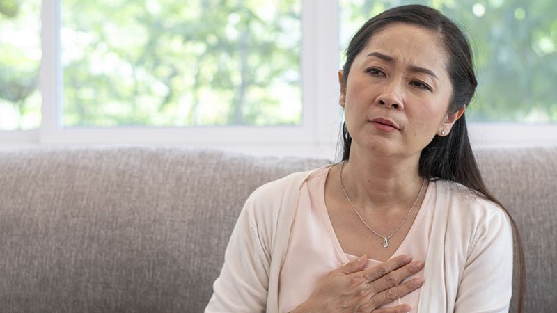 A mature woman with black hair holds her hand over her chest, with a sad or pained look on her face.