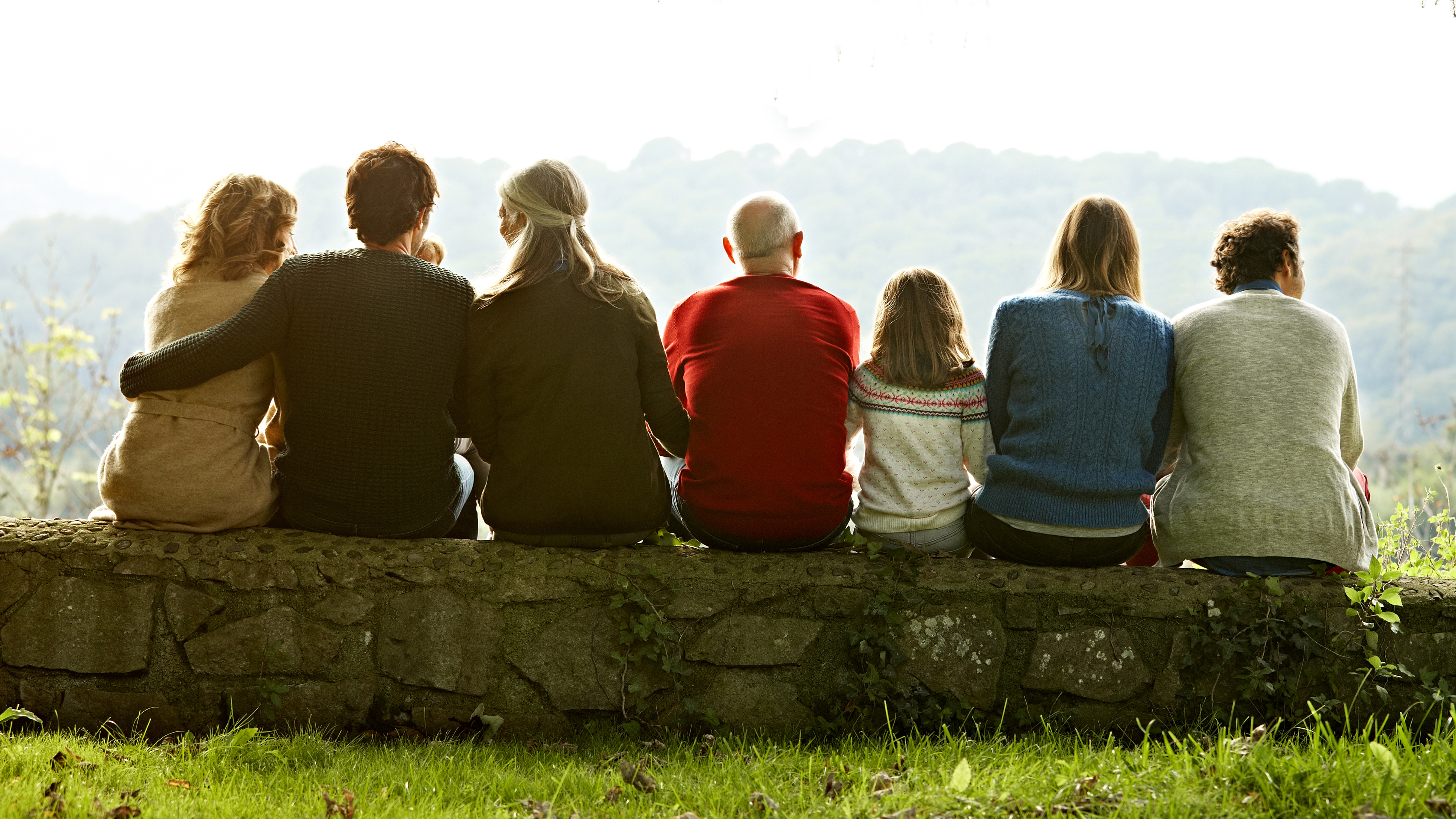 A multigenerational family viewed from behind sits in a row on a stone wall looking out over a valley.